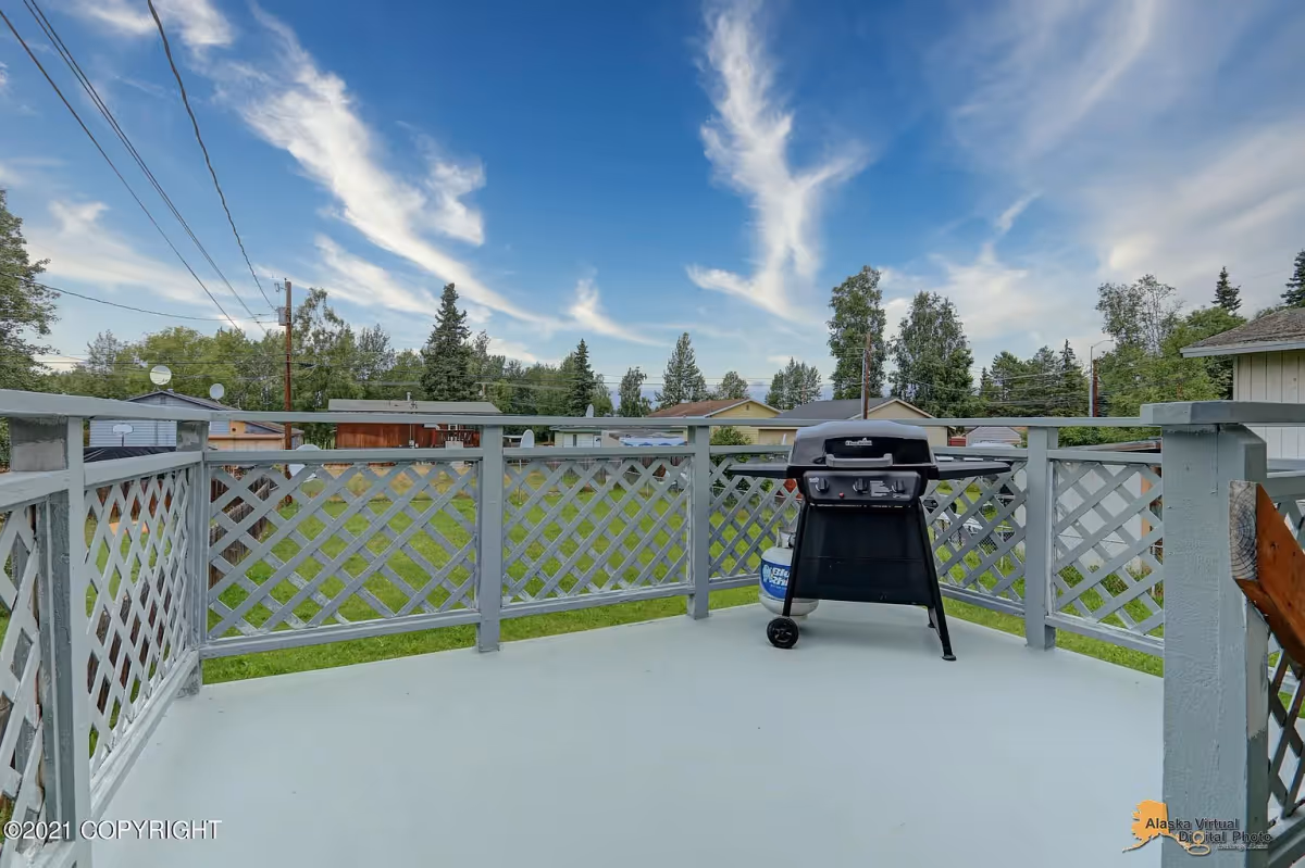Outdoor patio area with a gray lattice railing and a black propane grill. The patio overlooks a grassy backyard with trees and neighboring houses under a blue sky with scattered clouds.