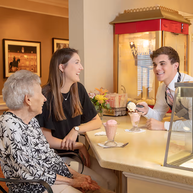 An elderly woman and a younger woman sit at a counter smiling while a young man scoops ice cream into sundaes in a communal dining area.