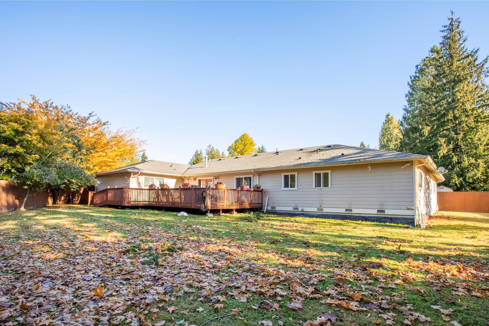 Exterior view of a single-story residential building with a wooden deck, surrounded by a grassy yard covered with fallen leaves and trees in the background under a clear blue sky.