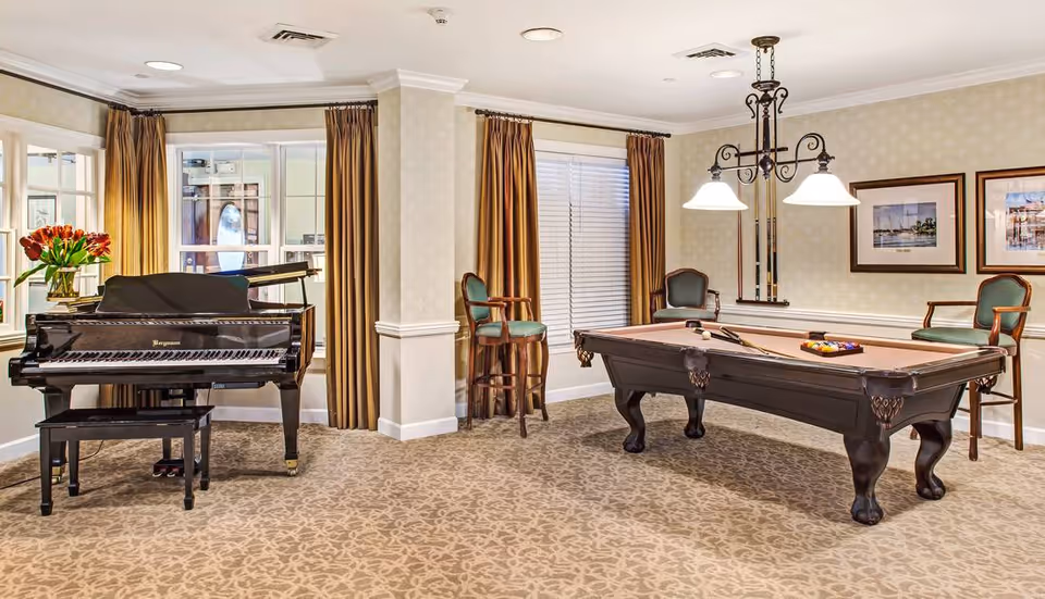 A well-lit recreational room featuring a grand piano with a vase of red flowers on top, a pool table with billiard balls and cues, three wooden chairs with green cushions, two framed pictures on the wall, and large windows with brown curtains.