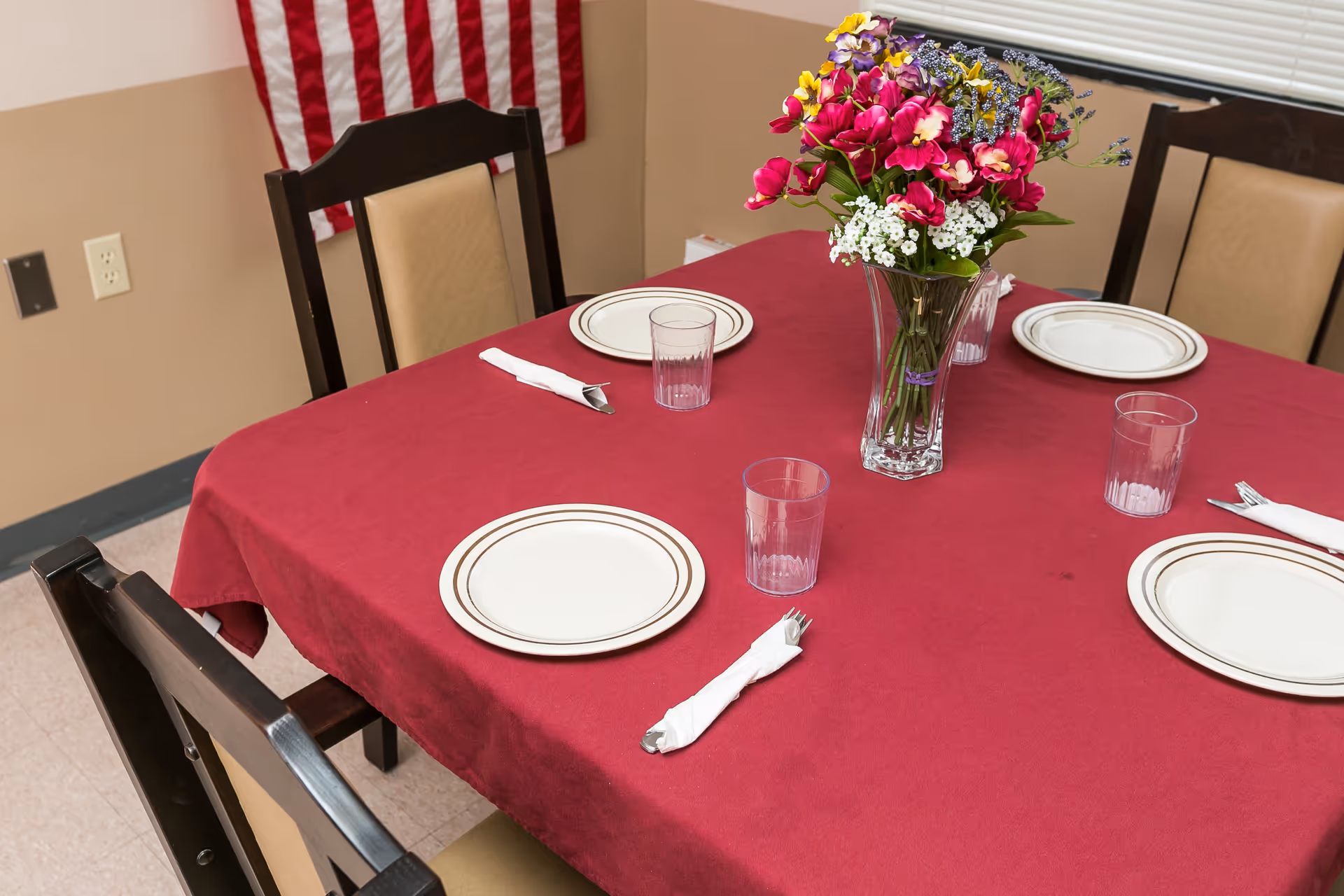A dining table covered with a red tablecloth set with plates, plastic cups, napkins, and a floral centerpiece, with chairs and an American flag on the wall.