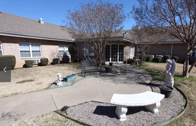 Outdoor courtyard with a paved walkway, white stone bench, decorative statues, trees and a one-story brick building with patio doors.