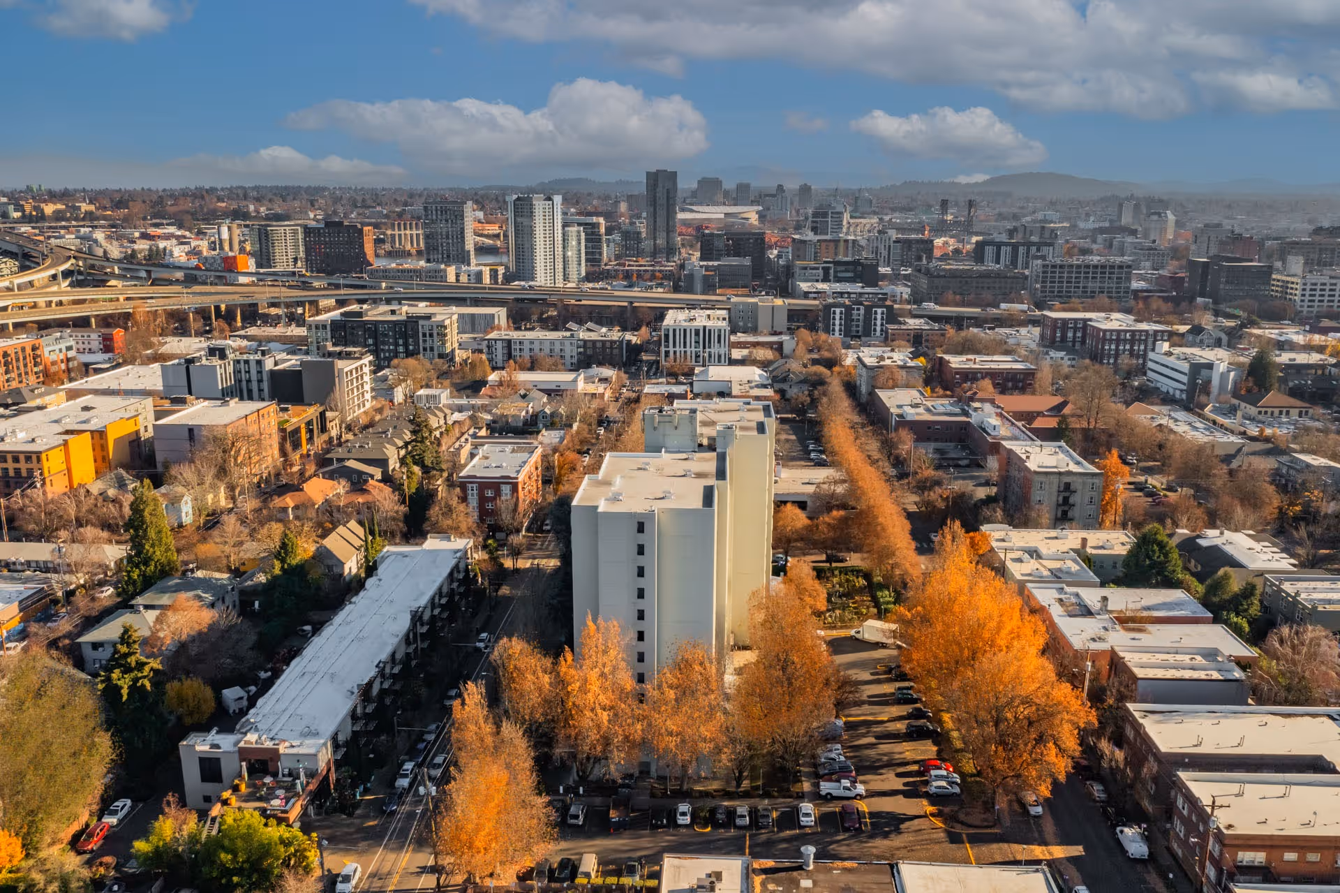 Aerial view of an urban area featuring a tall white building surrounded by trees with autumn foliage, parking lots, and various other buildings under a partly cloudy sky.