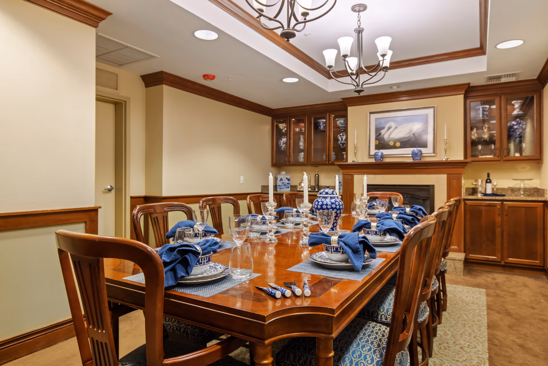A formal dining room with a long wooden table set with blue-and-white china, navy napkins, glassware, and chandeliers overhead.