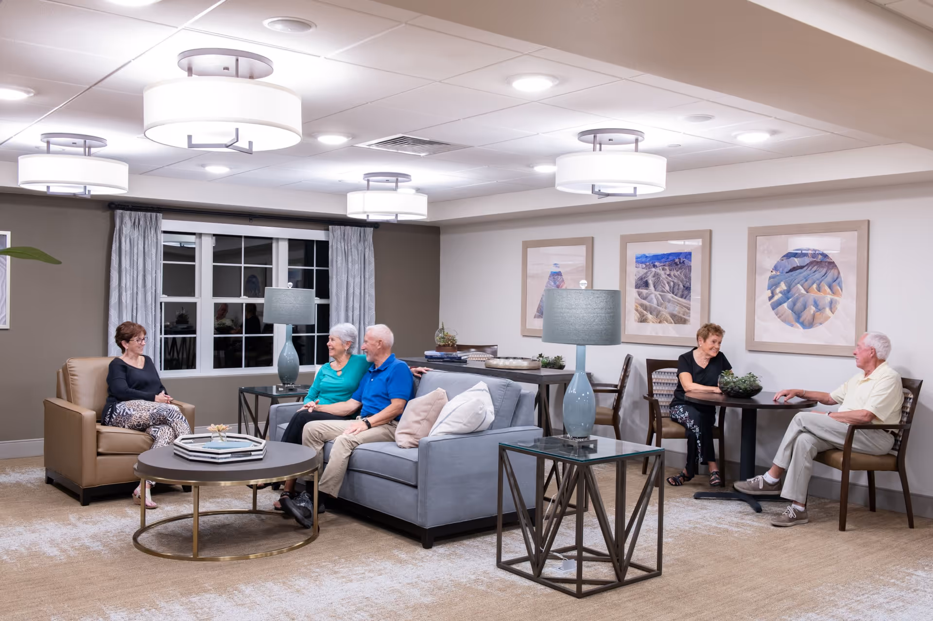 A well-lit senior living facility common area with five elderly people sitting and conversing. There are two seating areas: one with a gray sofa and a beige armchair around a round coffee table, and another with two chairs at a small table. The room has modern light fixtures on the ceiling, framed landscape artwork on the walls, and two blue table lamps on glass side tables.