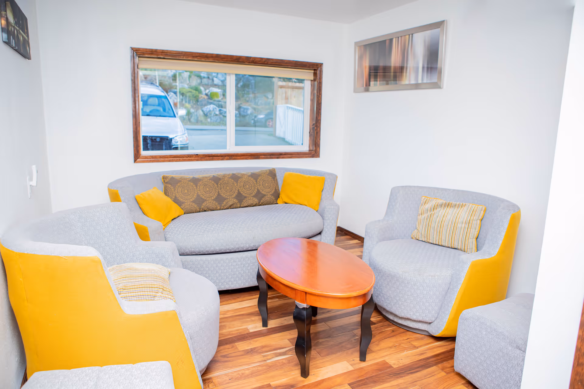 A small living room with a wooden floor, featuring a gray and yellow loveseat, two matching armchairs, and a wooden oval coffee table in the center. There is a window with a wooden frame showing a parked car outside, and a framed abstract artwork on the white wall.