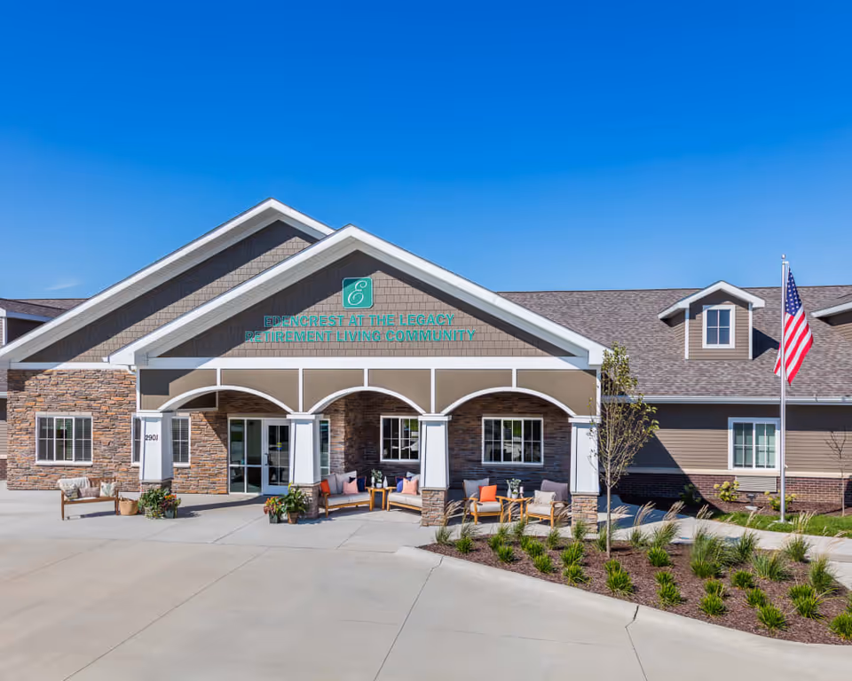 Front entrance of Edencrest at The Legacy retirement community building with stone facade, covered porch seating, and an American flag.