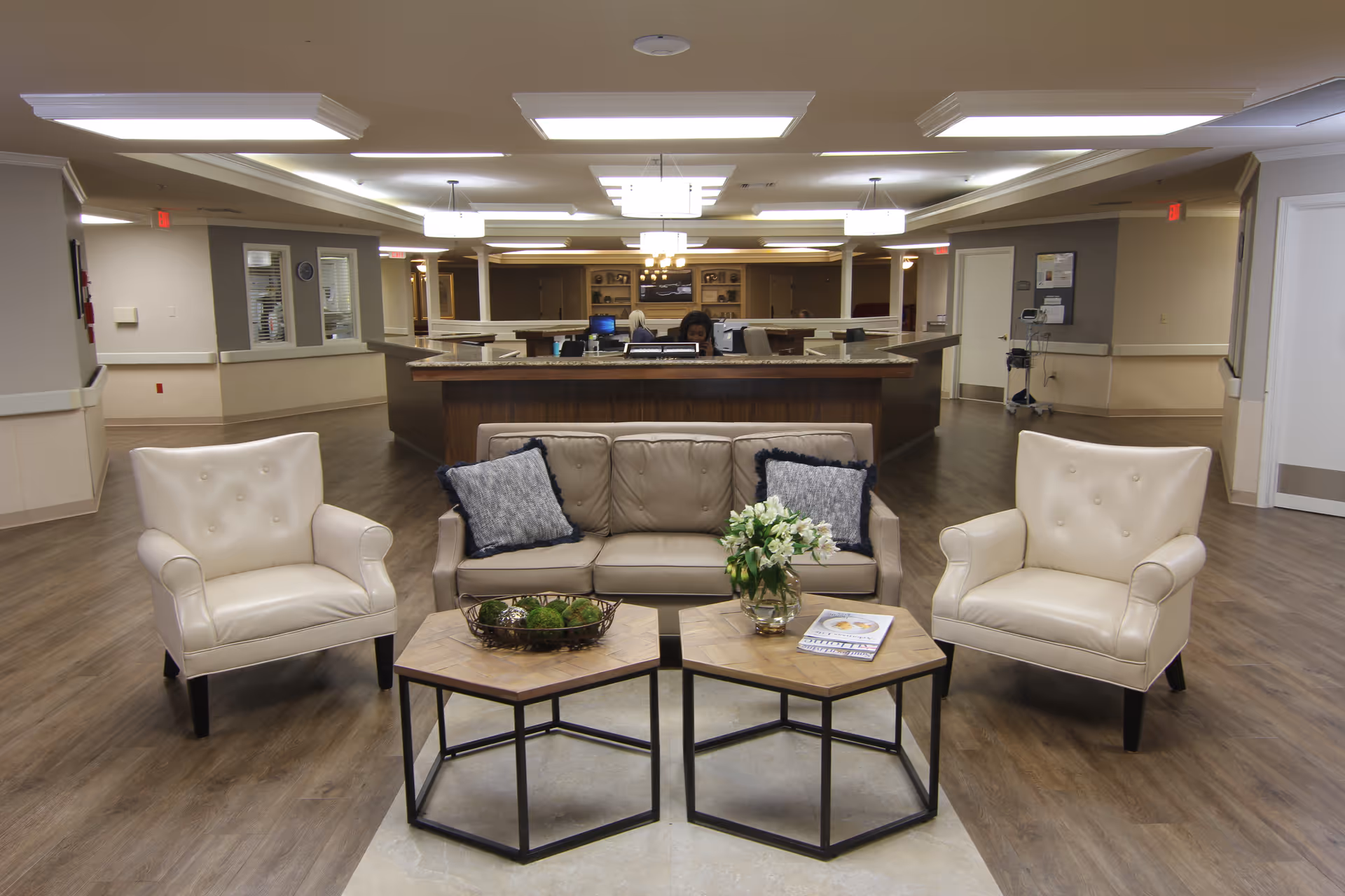 A nursing facility reception area with a beige leather sofa and two matching armchairs arranged around two hexagonal wooden coffee tables. The tables hold a decorative bowl with greenery and a vase with white flowers. Behind the seating area is a large reception desk with two staff members working. The space has wood flooring, neutral-colored walls, and modern ceiling lights.