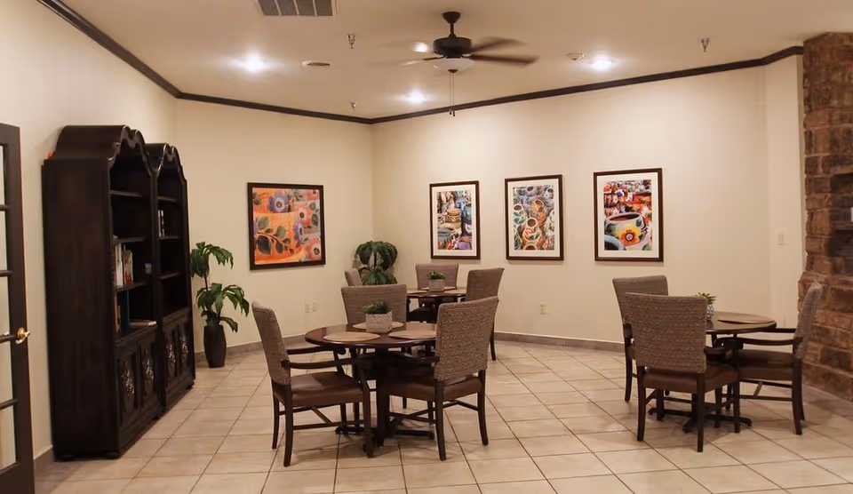 A well-lit room with several round tables and cushioned chairs arranged for seating. The walls are decorated with colorful framed artwork, and there is a dark wooden bookshelf on the left side. The floor is tiled, and a ceiling fan is mounted in the center of the ceiling. A stone wall is partially visible on the right side.
