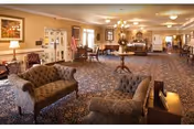 Spacious carpeted common lounge with upholstered sofas, chairs, small tables, chandeliers, and a reception/dining area in the background.