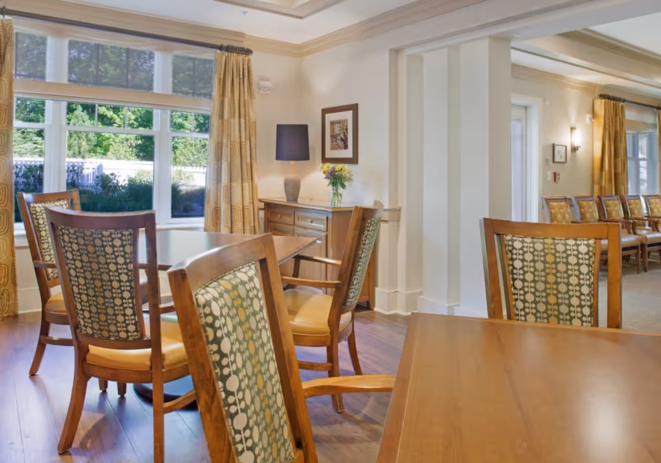 A bright and inviting dining room with wooden tables and chairs featuring patterned upholstery. Large windows with yellow curtains allow natural light to fill the room. A sideboard with a lamp and a vase of flowers is against the wall, and additional seating is visible in the background.