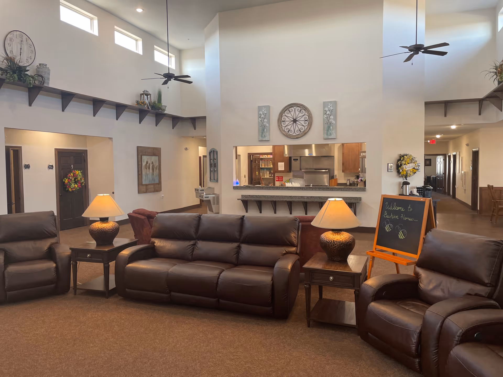 A spacious and well-lit common area in a senior living facility featuring brown leather sofas and armchairs arranged around two wooden side tables with lamps. The room has high ceilings with ceiling fans and small windows near the ceiling. A large clock and decorative wall art are mounted above a pass-through window to the kitchen area. A chalkboard sign with a welcome message and bee drawings is placed near the seating area. Hallways and doors are visible in the background.