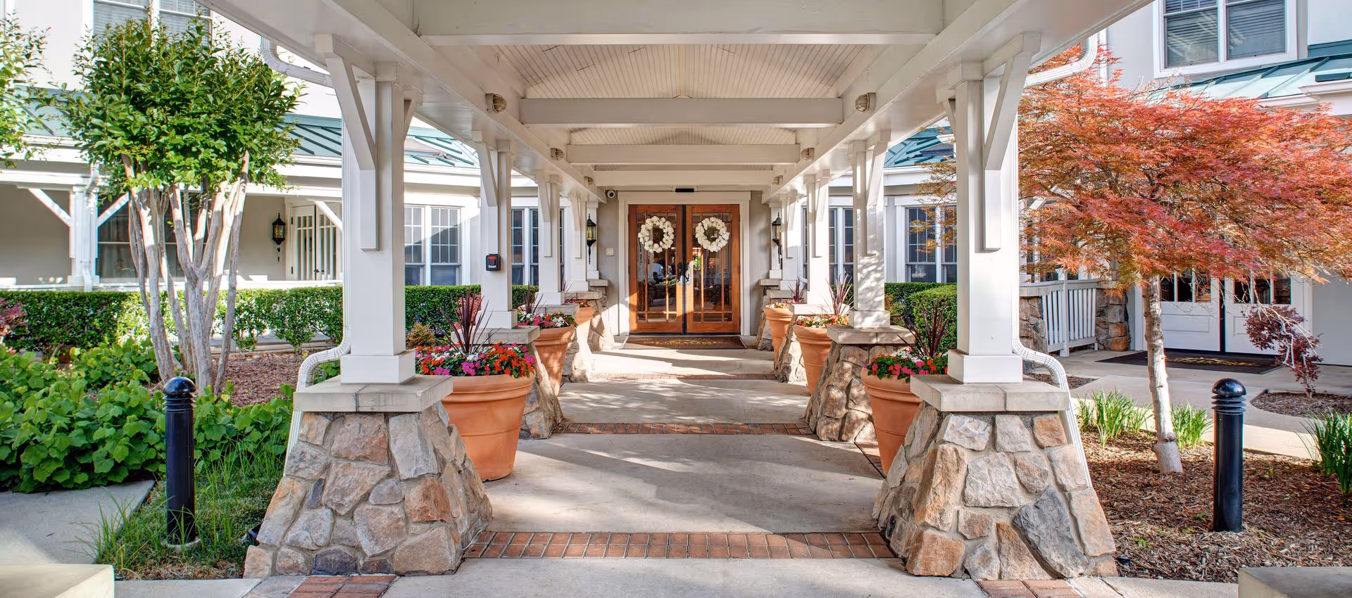 Covered entrance walkway to a building with stone pillars and large terracotta pots filled with colorful flowers on either side. The double wooden doors at the end of the walkway have white wreaths hanging on them. Surrounding the entrance are well-maintained shrubs and trees, including a red-leafed tree on the right.