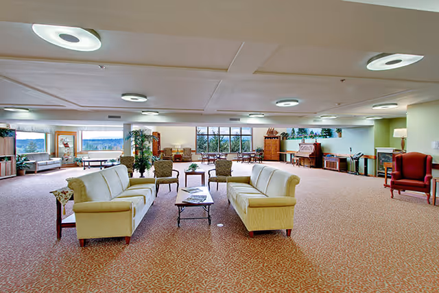 A spacious senior living facility common area with beige sofas arranged around a coffee table, several armchairs, large windows letting in natural light, a piano, and decorative plants. The room has patterned carpet and modern ceiling lights.