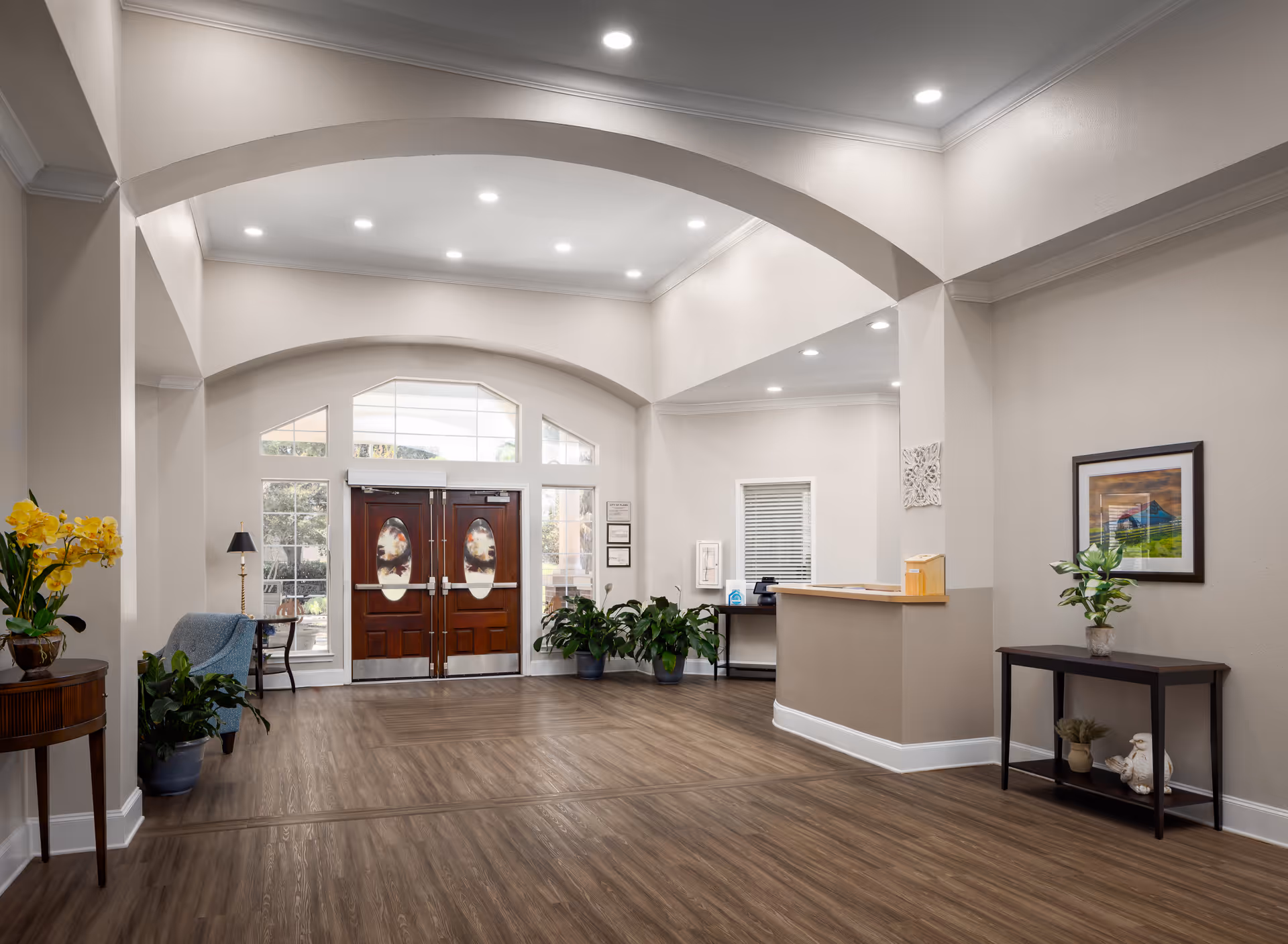 Spacious and well-lit reception area with wooden double doors featuring oval glass panels, light-colored walls, and wood flooring. The room includes a reception desk, potted plants, a small table with a lamp and chair, a side table with a vase of yellow flowers, and framed artwork on the wall.