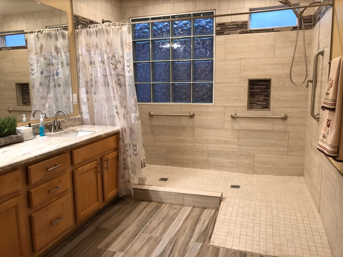 A bathroom with a large mirror above a wooden vanity with a marble countertop and a sink. There is a walk-in shower with a glass block window, grab bars, and a shower curtain with butterfly and floral patterns. The floor has wood-like tiles and the shower area has small square tiles.