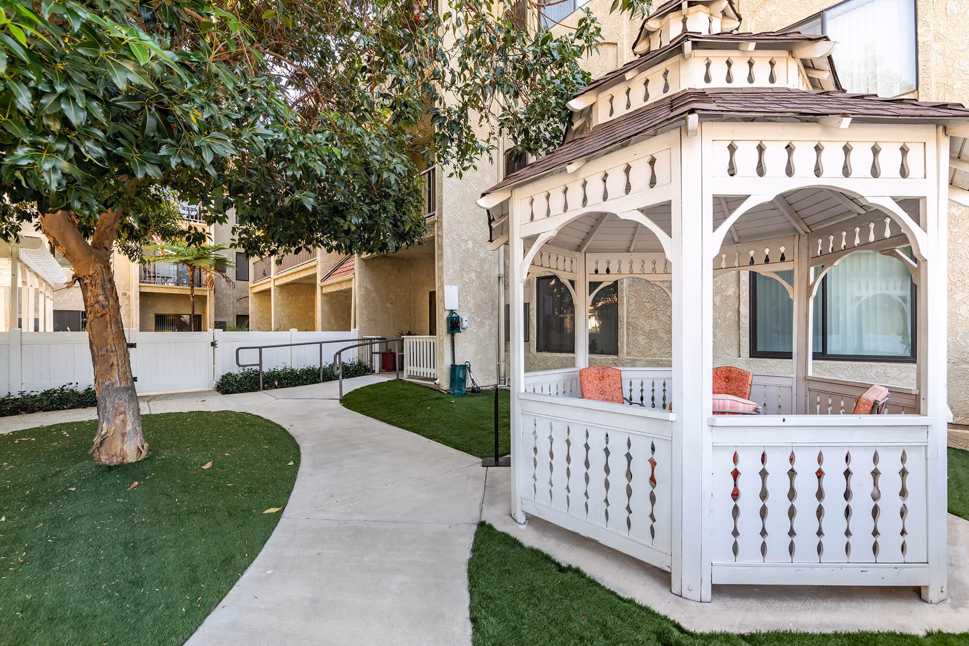 Outdoor area at Vista Corona Senior Living featuring a white wooden gazebo with cushioned chairs inside, a concrete pathway, green artificial grass, a tree, and surrounding buildings.