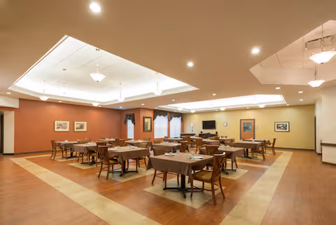 A spacious dining room with multiple tables covered with brown tablecloths and wooden chairs arranged neatly. The room has a warm color scheme with beige and brown tones, recessed lighting in the ceiling, and framed artwork on the walls. There is a television mounted on one wall and windows with curtains.