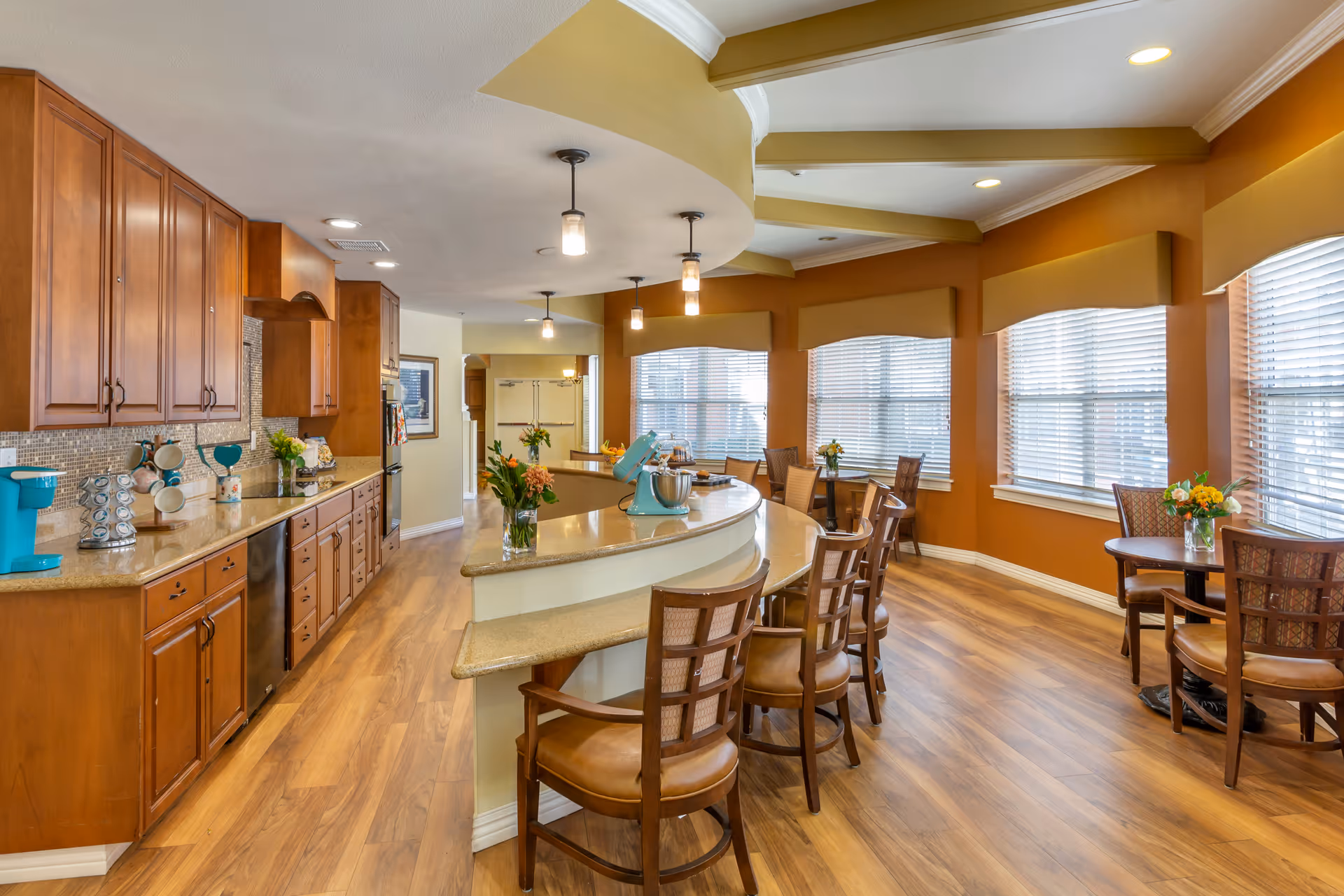 Open kitchen and dining area with a curved breakfast bar, wooden cabinets and chairs, small dining tables, and large windows.