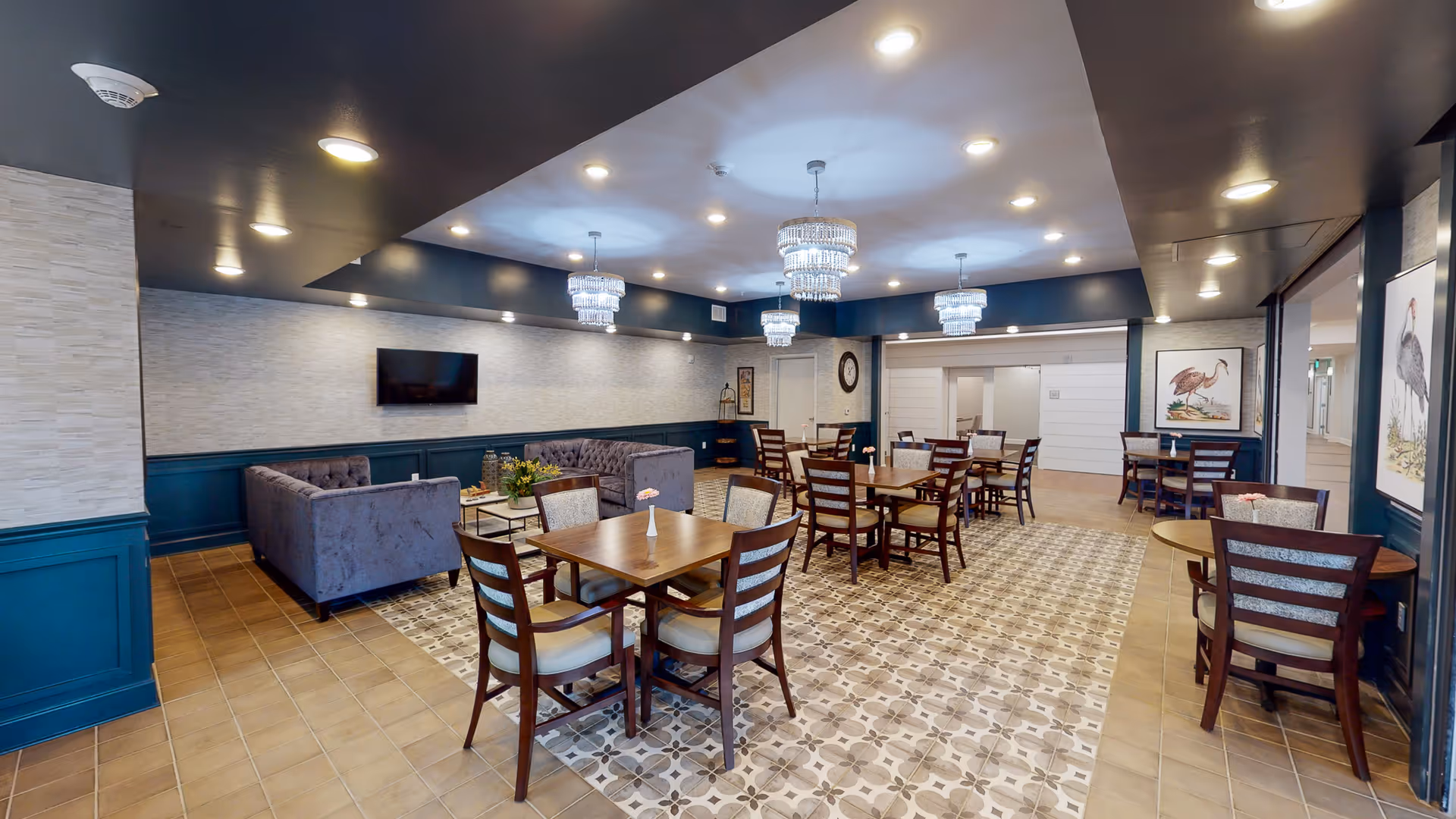 Open dining and lounge room with several wooden tables and chairs, tufted sofas, patterned tile floor, and hanging chandeliers.