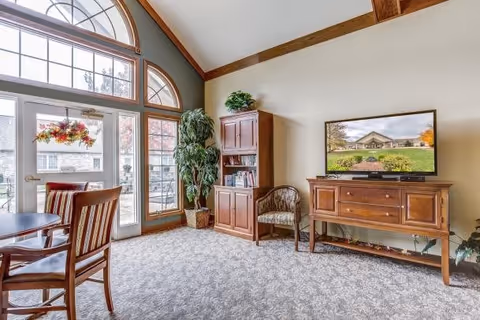 A bright and cozy living area with large arched windows and a glass door letting in natural light. The room features a wooden dining table with chairs, a tall wooden cabinet with books and decor, a potted plant, and a wooden TV stand with a flat-screen television displaying an image of a building and garden.