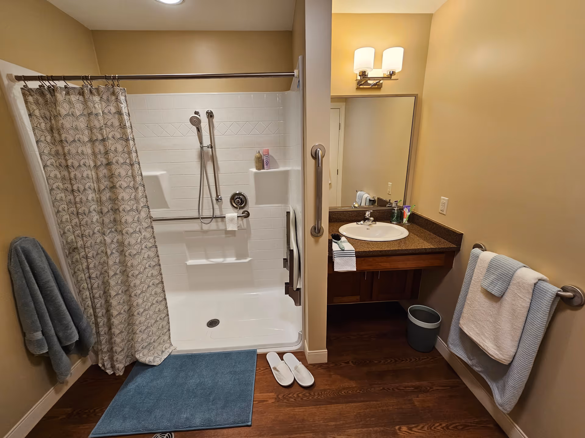 A bathroom with a walk-in shower featuring a patterned shower curtain, a handheld showerhead, and built-in shelves holding toiletries. There is a blue bath mat on the wooden floor and white slippers placed nearby. The sink area has a brown countertop with a mirror above it, a wall-mounted light fixture, and towels hanging on racks on both sides of the room.