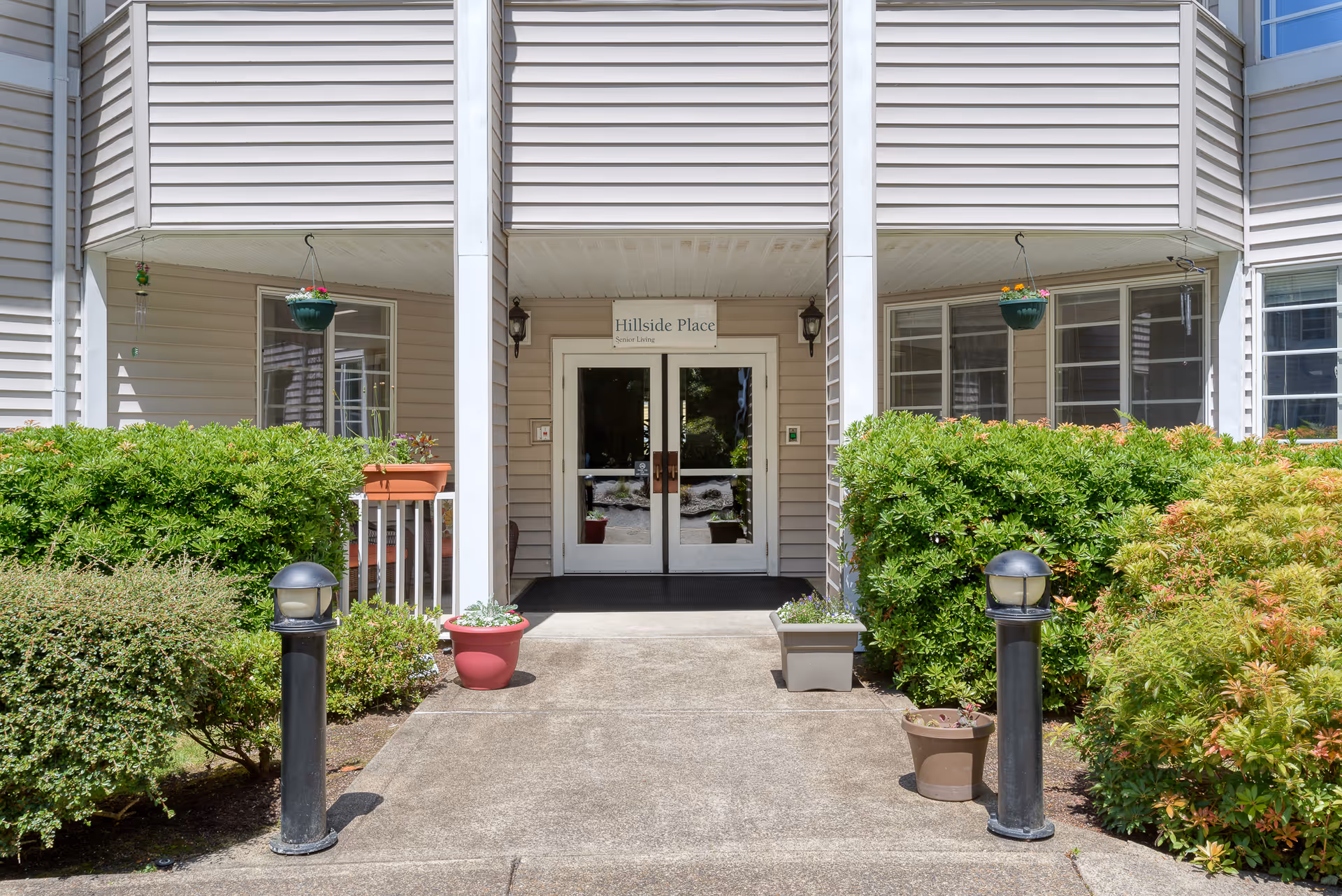 Entrance to Hillside Place Senior Living facility with double glass doors, surrounded by beige siding and white columns. There are green bushes and potted plants on either side of the concrete walkway leading to the doors. Two black outdoor lamp posts are positioned along the walkway.