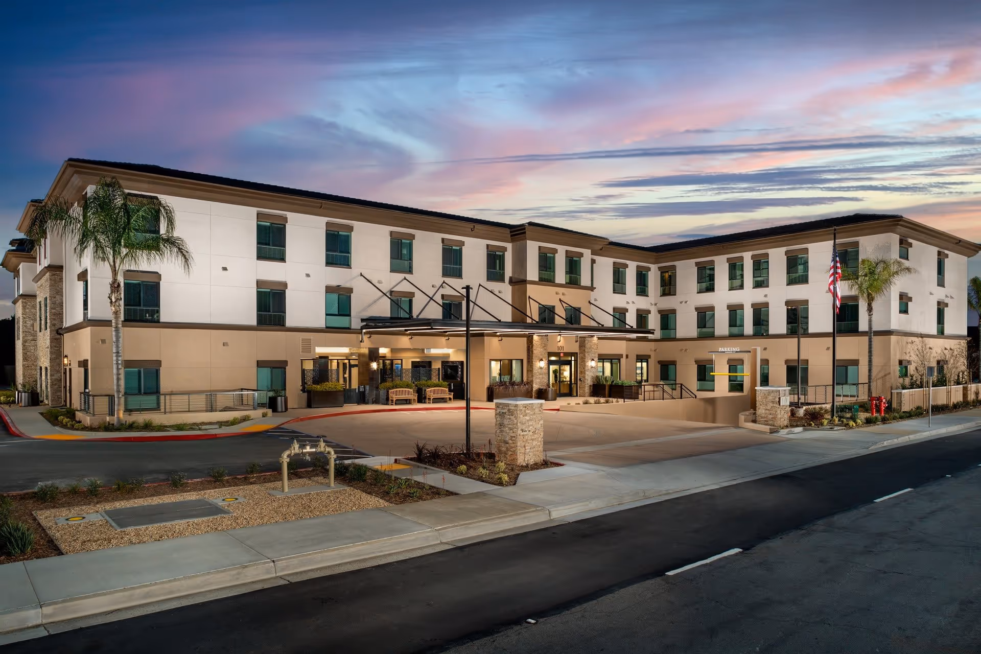 Exterior view of a modern three-story senior living facility building at dusk with a clear sky showing pink and blue hues. The building has multiple windows, a covered entrance with benches, palm trees, and an American flag near the entrance. The surrounding area includes a driveway, sidewalk, and landscaped sections.