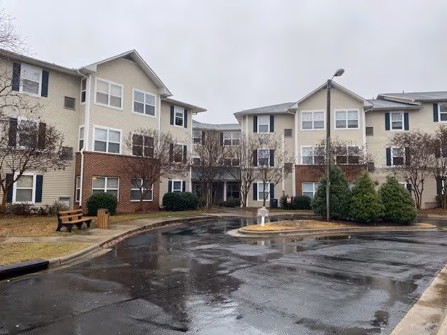 Exterior view of a multi-story senior living facility with beige siding and brick accents. The building is surrounded by leafless trees, shrubs, and a wet driveway with a small roundabout. A bench and trash can are visible on the left side near the sidewalk.