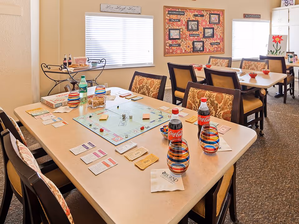 A bright communal dining/activity room with tables and chairs set up for a Monopoly game, colorful glasses, Coca-Cola bottles, and wall decorations.
