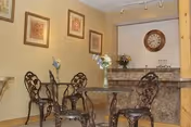 A small dining area with a glass-top table and four ornate metal chairs. The walls are decorated with framed artwork, and there is a clock and flower arrangements on a granite countertop in the background.