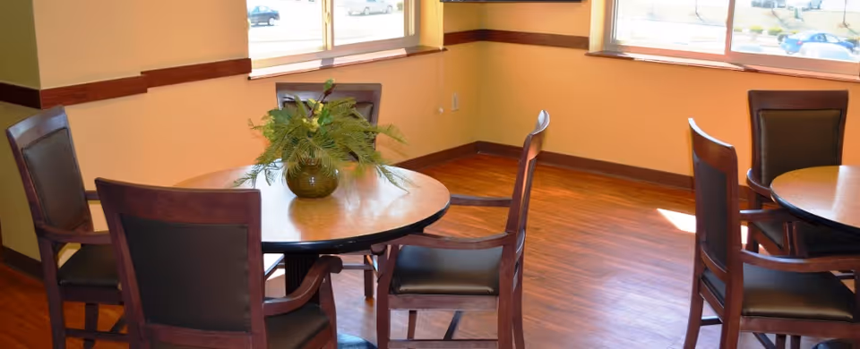 Small communal dining area with round wooden tables, chairs, a potted plant centerpiece, and windows letting in daylight.