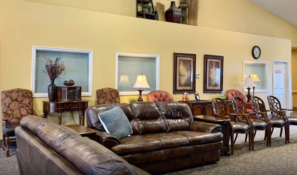 A cozy living room area with two brown leather sofas, several wooden chairs with cushioned seats, and side tables with lamps. The walls are painted yellow and decorated with framed artwork, a clock, and decorative vases on a high shelf. There are windows with blinds and a carpeted floor with a subtle pattern.