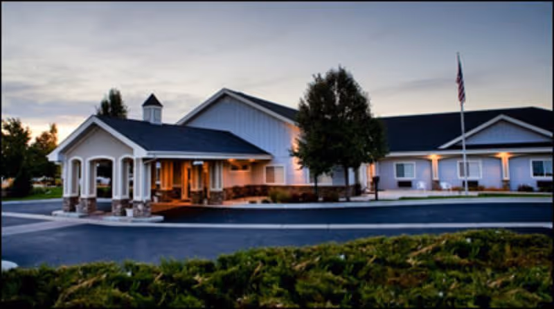 Exterior view of a single-story senior living facility building at dusk with a covered entrance, well-maintained landscaping, a tree, and an American flag on a flagpole.