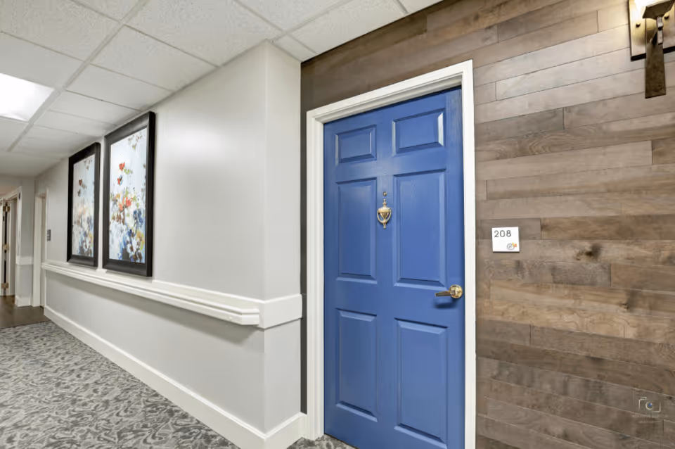 Interior hallway of a senior living facility with a blue door labeled 208 on the right side. The hallway has light-colored walls with a white chair rail, patterned carpet flooring, and two framed floral artworks on the left wall. The wall around the door is covered with wooden panels.
