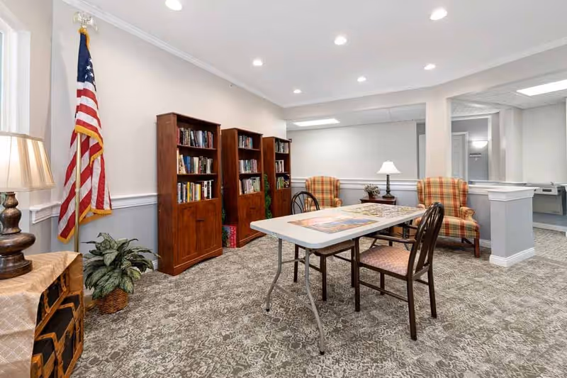 A cozy interior room with a patterned carpet, three wooden bookshelves filled with books, a folding table with two chairs, two plaid upholstered armchairs, a table lamp on a small side table, an American flag in the corner, and a potted plant near a window with a lamp on a wooden stand.