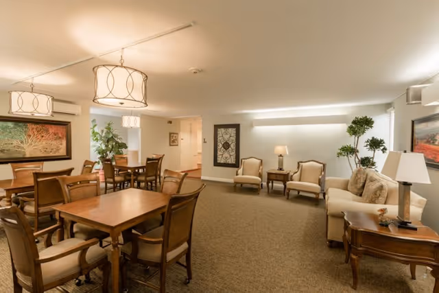 A well-lit common area in a senior living facility featuring multiple wooden tables with chairs, a seating area with two armchairs and a sofa, decorative plants, framed artwork on the walls, and ceiling light fixtures.