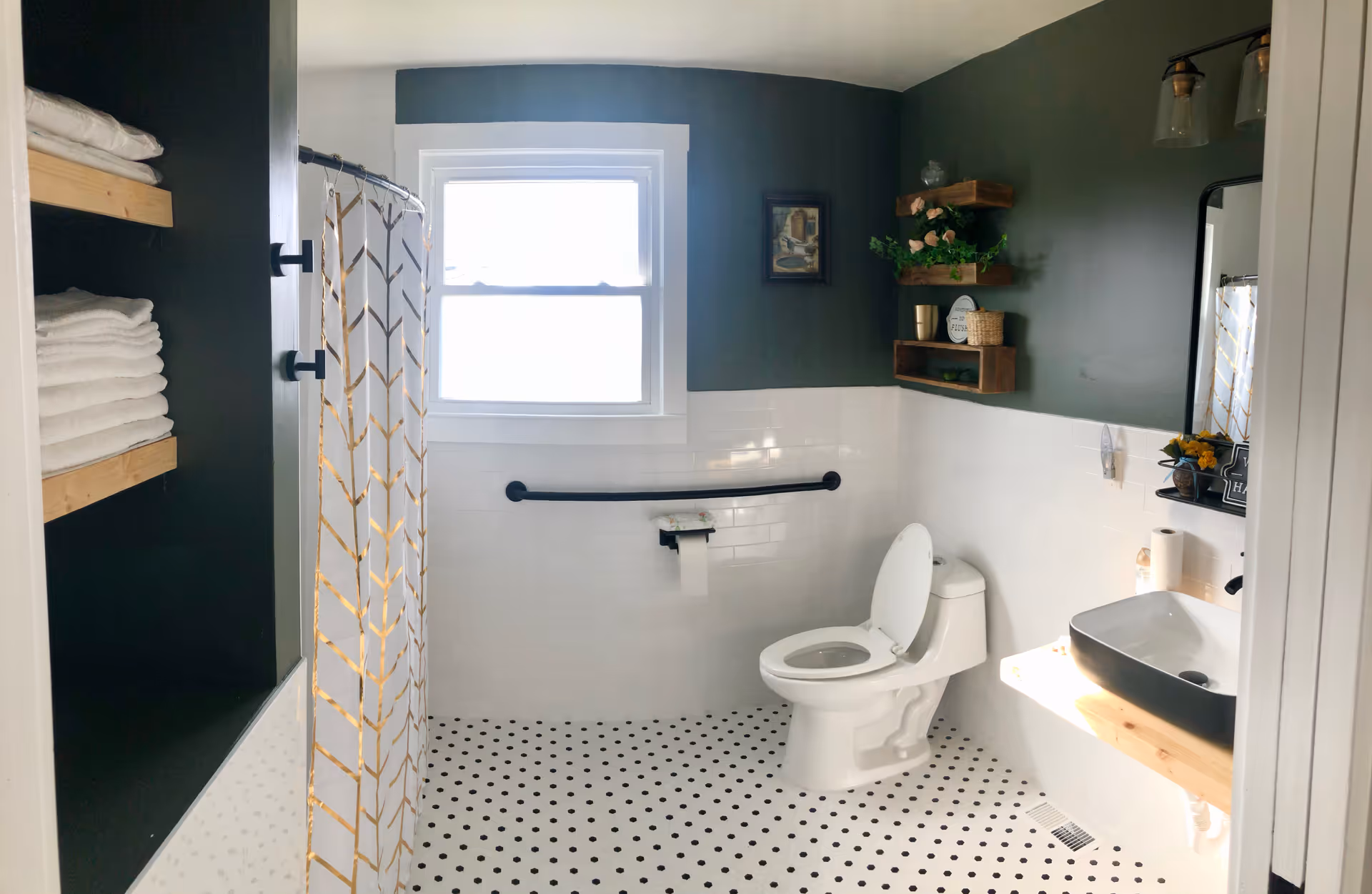 A clean and modern bathroom with white and black polka dot floor tiles, white tiled walls halfway up, and dark green paint above. There is a white toilet with the lid open, a black grab bar with a toilet paper holder, a white sink with a black basin on a wooden countertop, and a mirror above the sink. Shelves with decorative items and towels are mounted on the walls. A window lets in natural light, and a shower curtain with a geometric pattern is visible on the left side.