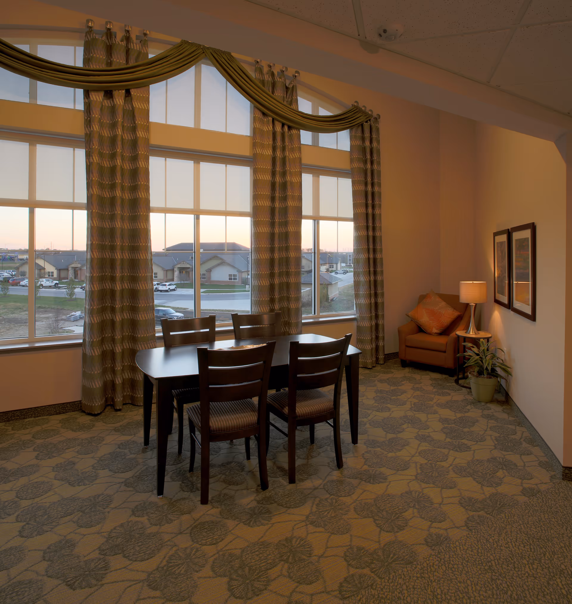 A cozy interior room with a dark wooden table and four matching chairs placed near large windows with patterned curtains. The windows offer a view of houses and cars outside. In the corner, there is a comfortable armchair with a decorative pillow, a small side table with a lamp, two framed pictures on the wall, and a potted plant on the floor.