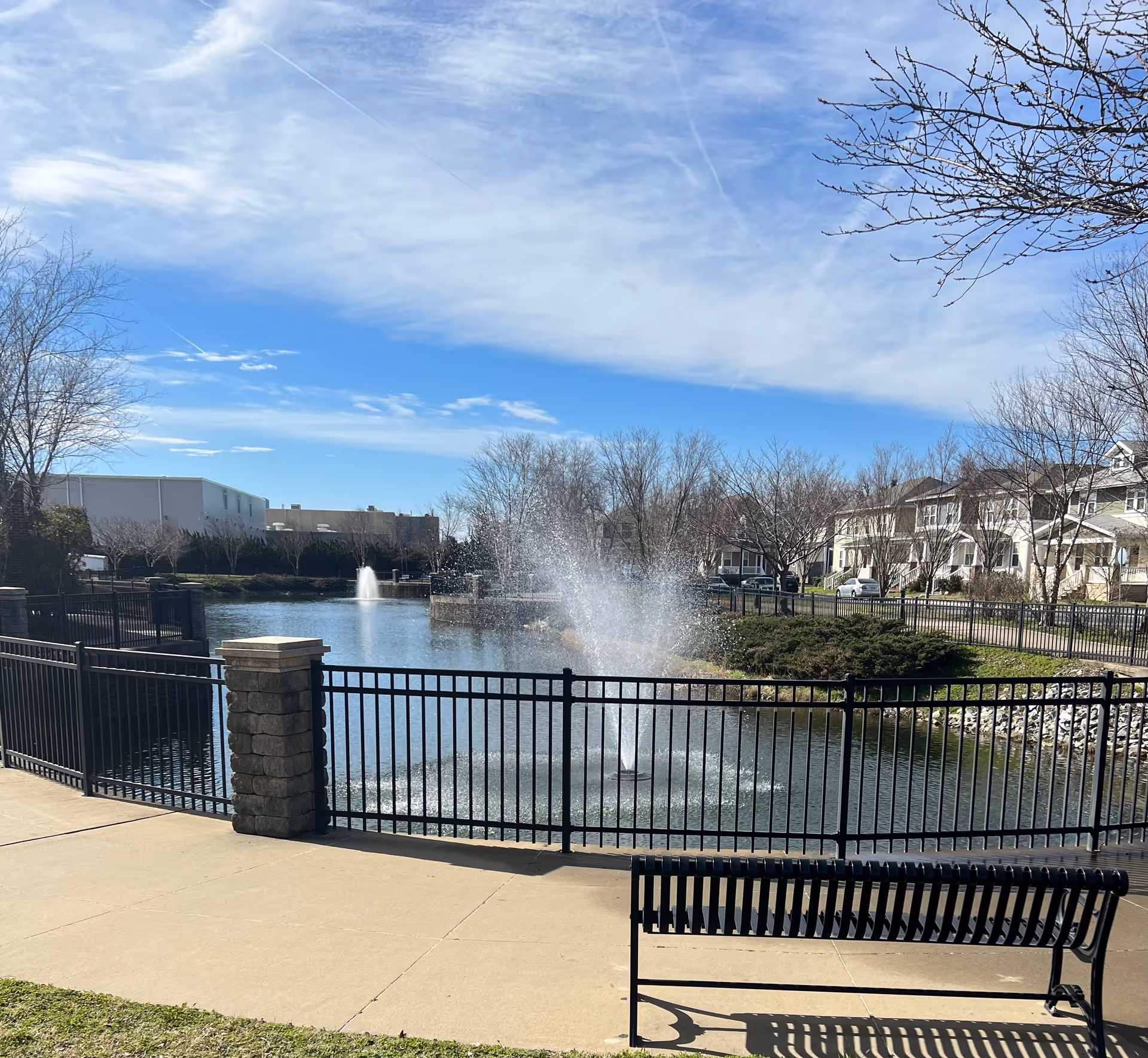 Outdoor scene featuring a pond with two water fountains surrounded by a black metal fence. There is a black metal bench on a concrete walkway in the foreground. Leafless trees and residential buildings are visible in the background under a partly cloudy blue sky.