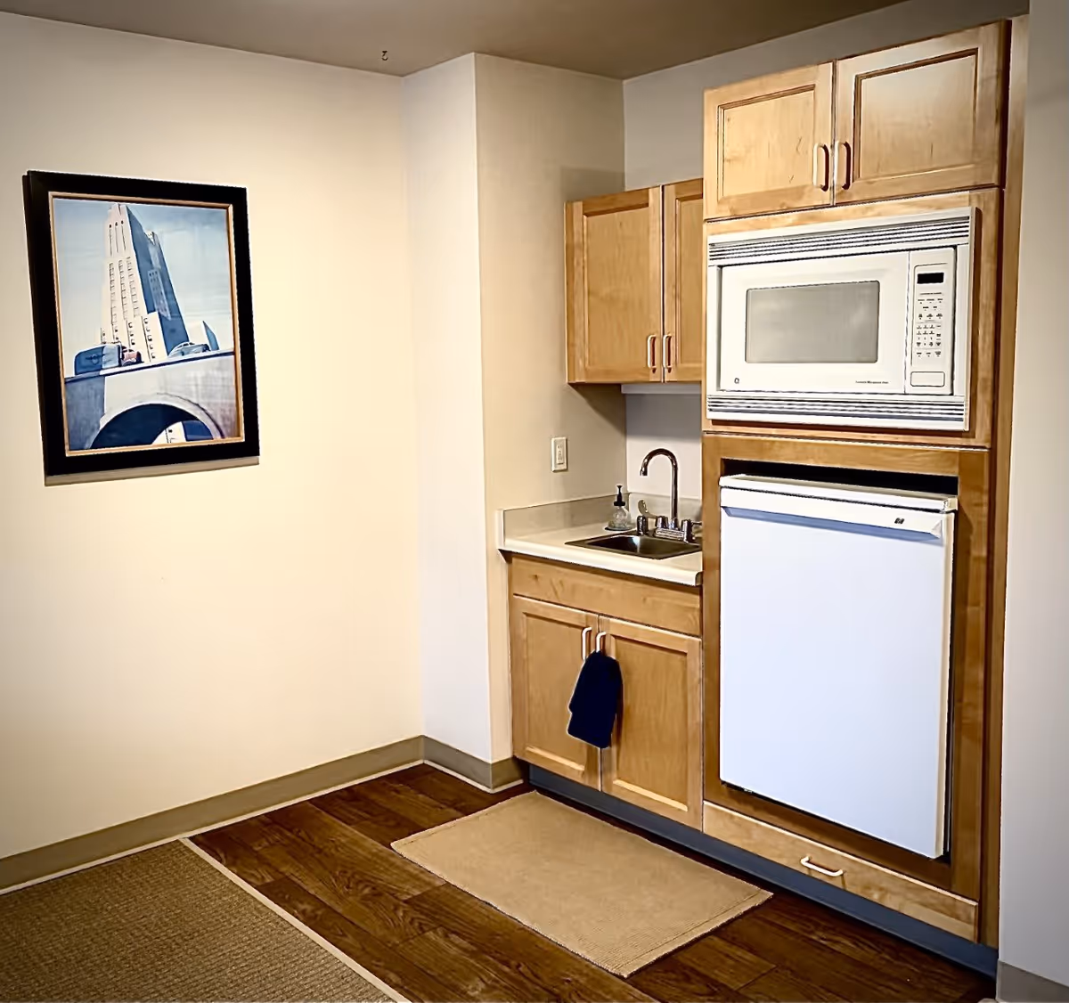 Small kitchenette area with light wood cabinets, a white microwave above a white mini refrigerator, a small sink with a faucet, and a dark towel hanging on the cabinet door. There is a framed picture of a building on the adjacent wall and a beige rug on the wooden floor.