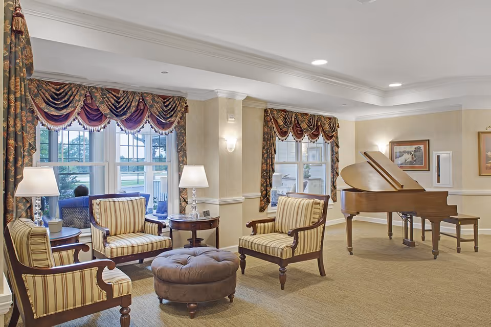 A cozy living room area with three striped armchairs arranged around a round tufted ottoman. Two side tables with lamps are placed near the chairs. Large windows with ornate drapes allow natural light to fill the room. A grand piano with a matching bench is positioned against the wall, and framed artwork hangs above it. The room has beige walls and carpeted flooring.