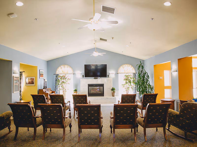 A bright and spacious common area with rows of wooden chairs facing a fireplace and a mounted flat-screen TV. The room has light blue walls, two large arched windows with curtains, ceiling fans, and several potted plants. There are doorways on either side leading to other rooms painted in warm yellow tones.