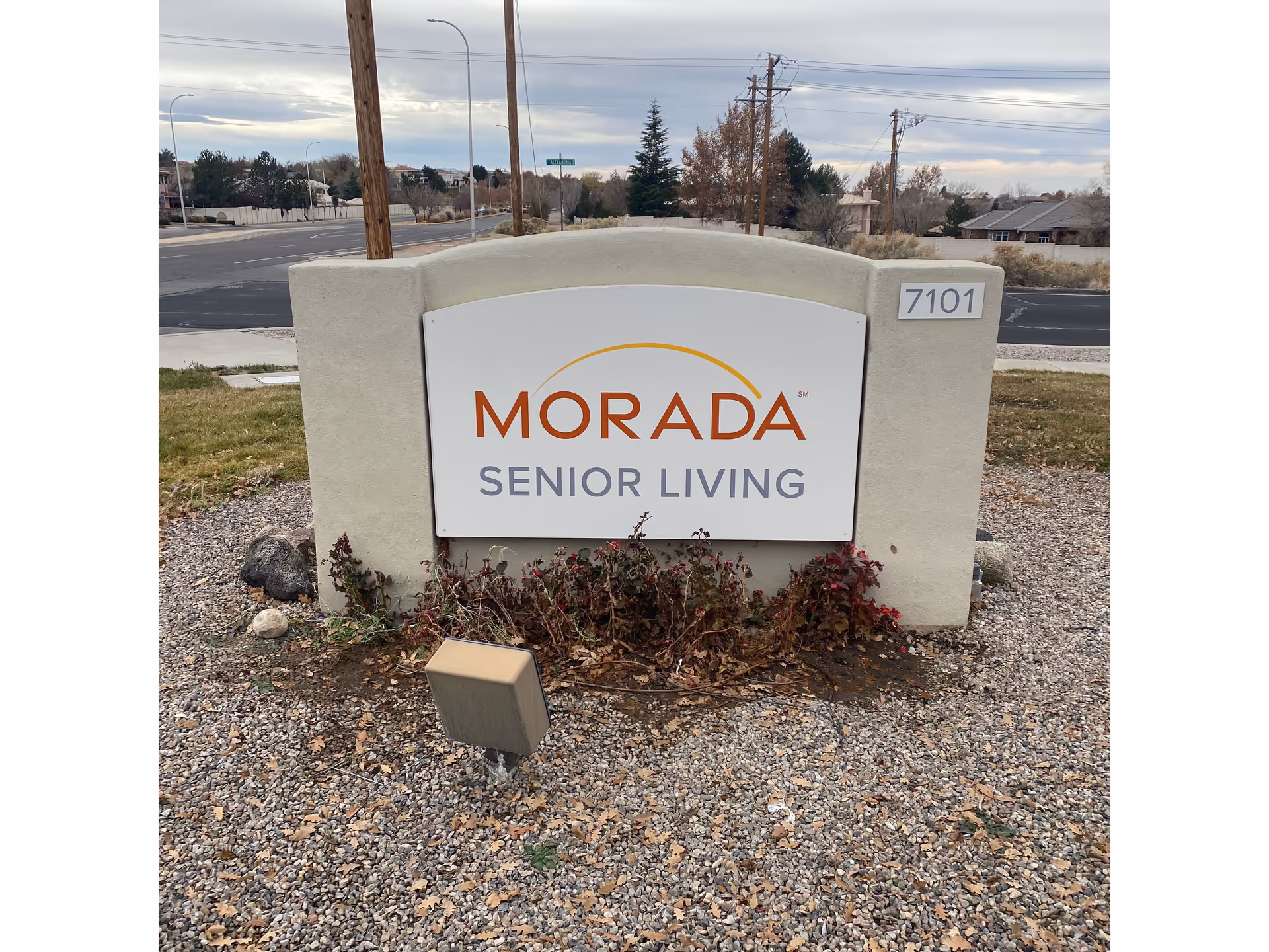 Outdoor sign for Morada Senior Living with the address number 7101 displayed on a white and beige stone structure, surrounded by gravel and some dried plants, with a street and trees in the background under a cloudy sky.