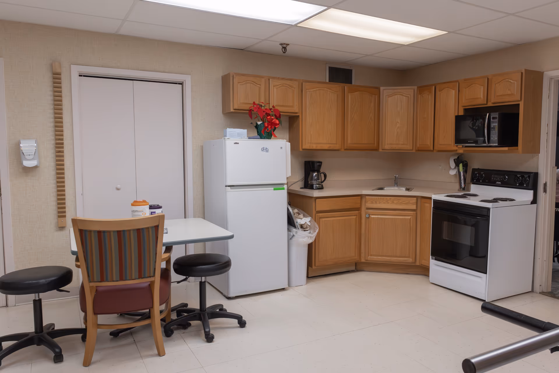 A small kitchen area with wooden cabinets, a white refrigerator, a white electric stove with an oven, a microwave, and a coffee maker. There is a table with three chairs, two black stools and one wooden chair with a striped backrest. A red poinsettia plant is on top of the refrigerator. The floor is tiled and the walls are beige.