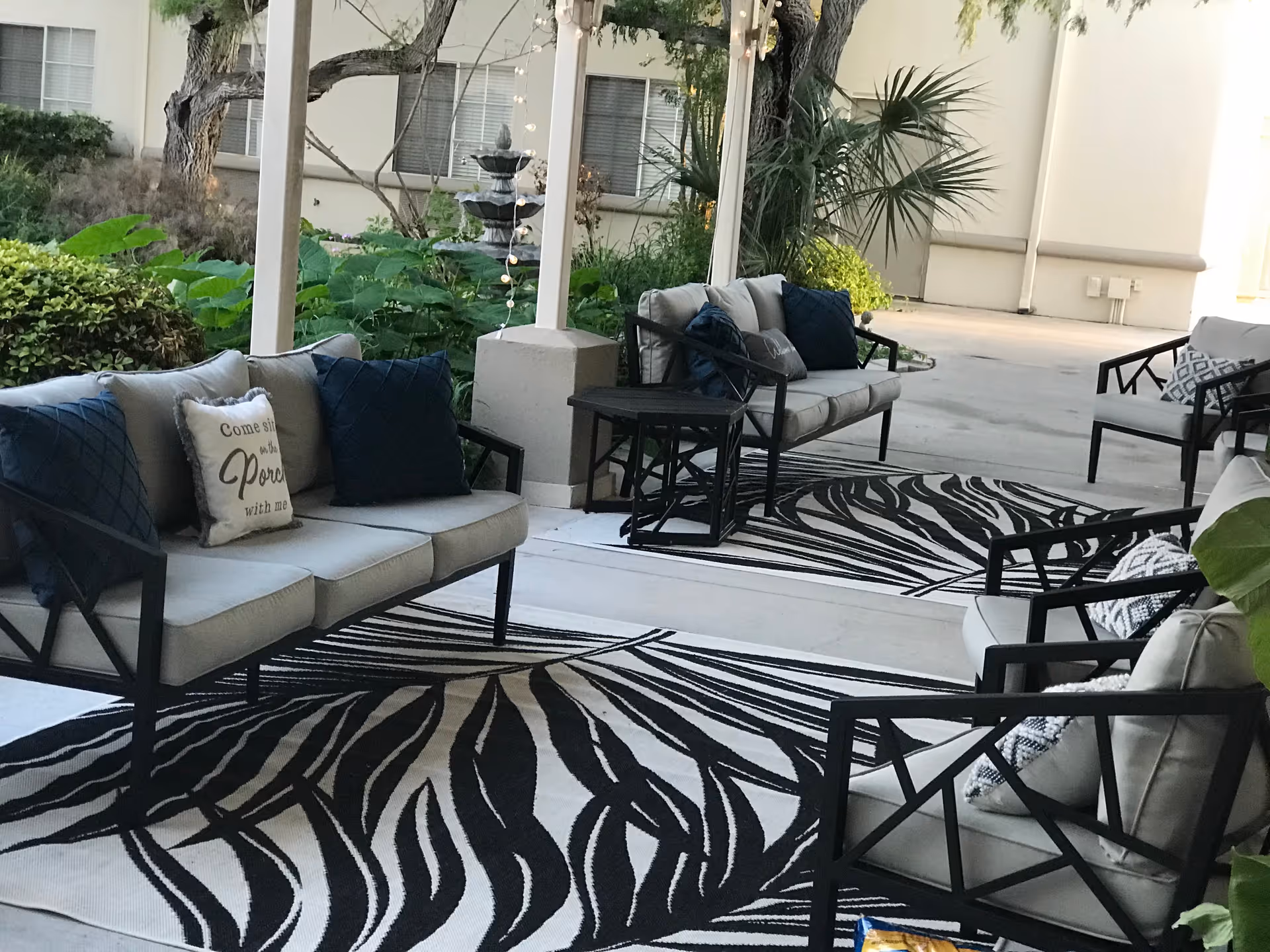 Outdoor covered patio area with cushioned seating including sofas and chairs arranged around black and white patterned rugs. There are decorative pillows on the furniture, some with text, and greenery including plants and trees surrounding the space.