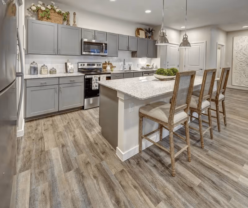Modern kitchen with gray cabinets, stainless steel appliances including a refrigerator, oven, and microwave. A large island with a granite countertop has three wooden bar stools with cushioned seats. The floor is wood-style laminate, and there are two pendant lights hanging above the island. Decorative items are placed on top of the cabinets and on the countertop.