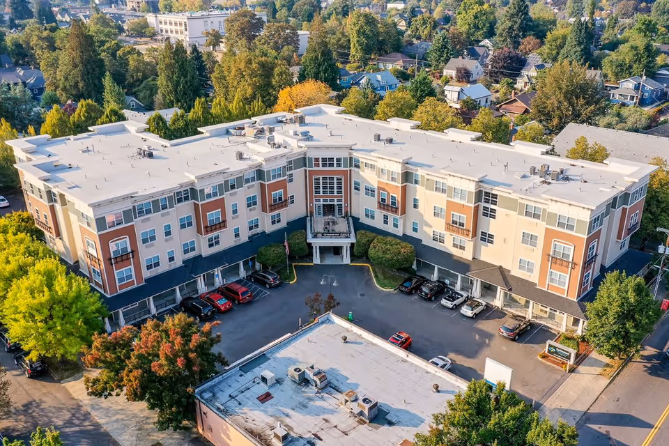 Aerial view of Sellwood Senior Living, a large multi-story residential building with a white roof, surrounded by trees and parked cars in the driveway. The building is situated in a neighborhood with many houses and greenery.