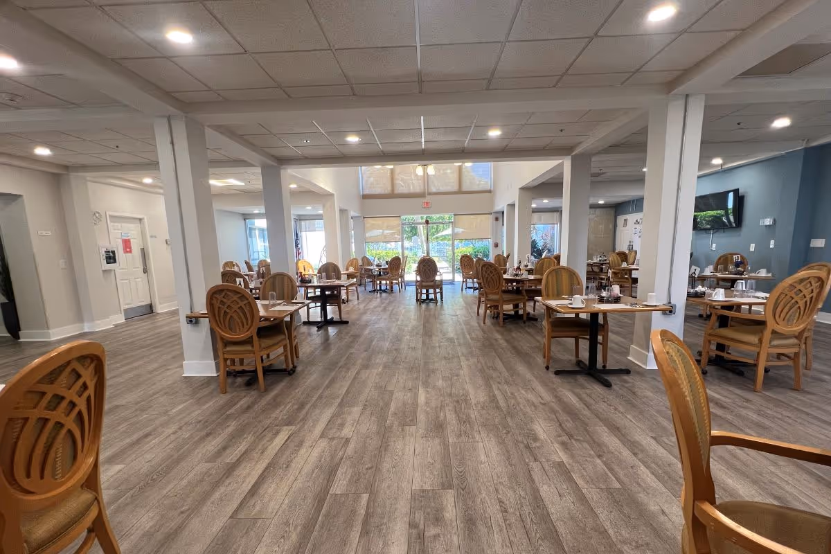 A spacious dining room with wooden tables and chairs arranged neatly. The room has large windows at the far end letting in natural light, a light-colored ceiling with recessed lighting, and wood-patterned flooring. Tables are set with cups and utensils, and a TV is mounted on the right wall.