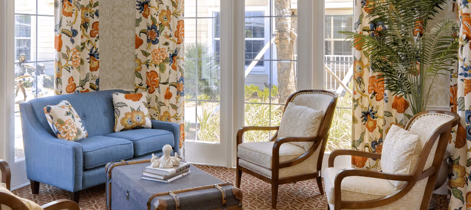 A cozy living room area with a blue loveseat adorned with floral pillows, two cushioned wooden armchairs, a vintage trunk used as a coffee table with decorative items on top, floral patterned curtains, and large windows letting in natural light with a view of greenery outside.